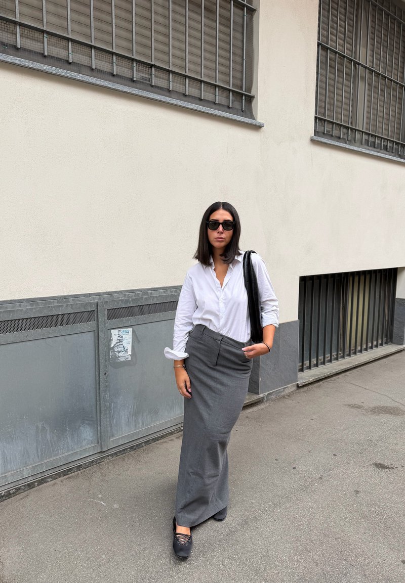 Camisa branca de botões, saia longa cinza, bolsa preta e óculos de sol. Tecido suave com corte ajustado; está de pé sobre uma superfície pavimentada perto de um edifício.