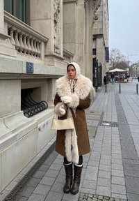 Femme en manteau marron avec grand col en fourrure, bonnet en laine, mitaines et bottes, debout sur le trottoir de la ville près d'un bâtiment en pierre.
