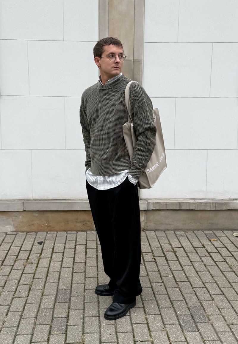 Olijfgroene gebreide trui met hoge kraag over een wit geruit shirt; zwarte wijde broek; beige handtas; zwarte schoenen; neutrale achtergrond.