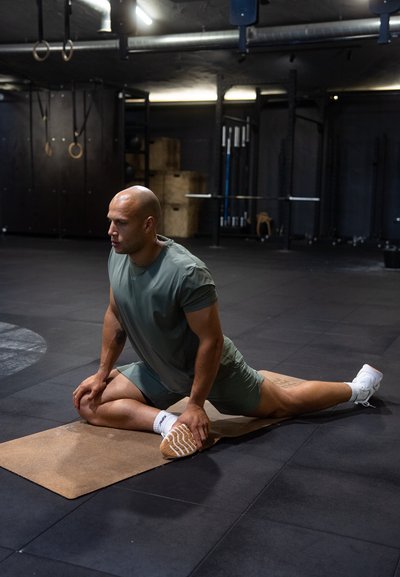 Persona realizando un ejercicio de estiramiento sobre una esterilla marrón, vestida con ropa deportiva verde oliva y zapatos blancos, en un suelo oscuro de gimnasio.