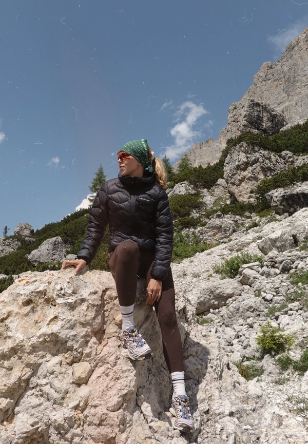 Melna pūkaina jaka, zaļa izšūta bandana, brūnas legingi, balti zeķes un daudzkrāsaini pārgājienu apavi uz akmeņainas virsmas zem zilas debess.