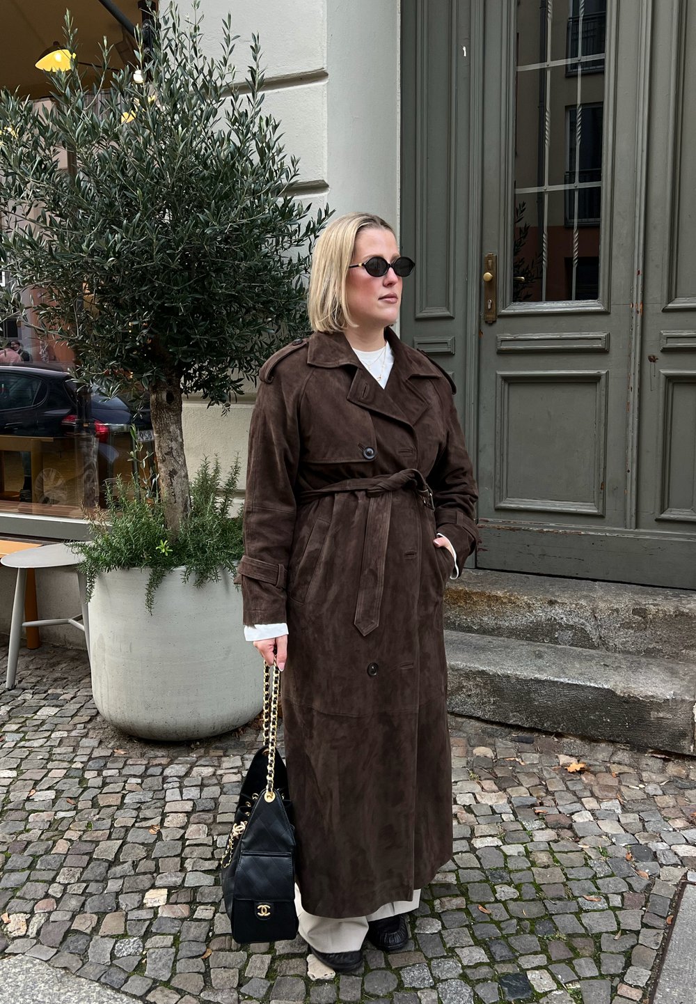 Femme portant un long trench-coat marron foncé, des lunettes de soleil, tenant un sac à main noir, debout sur une rue pavée près d'une grande plante en pot.