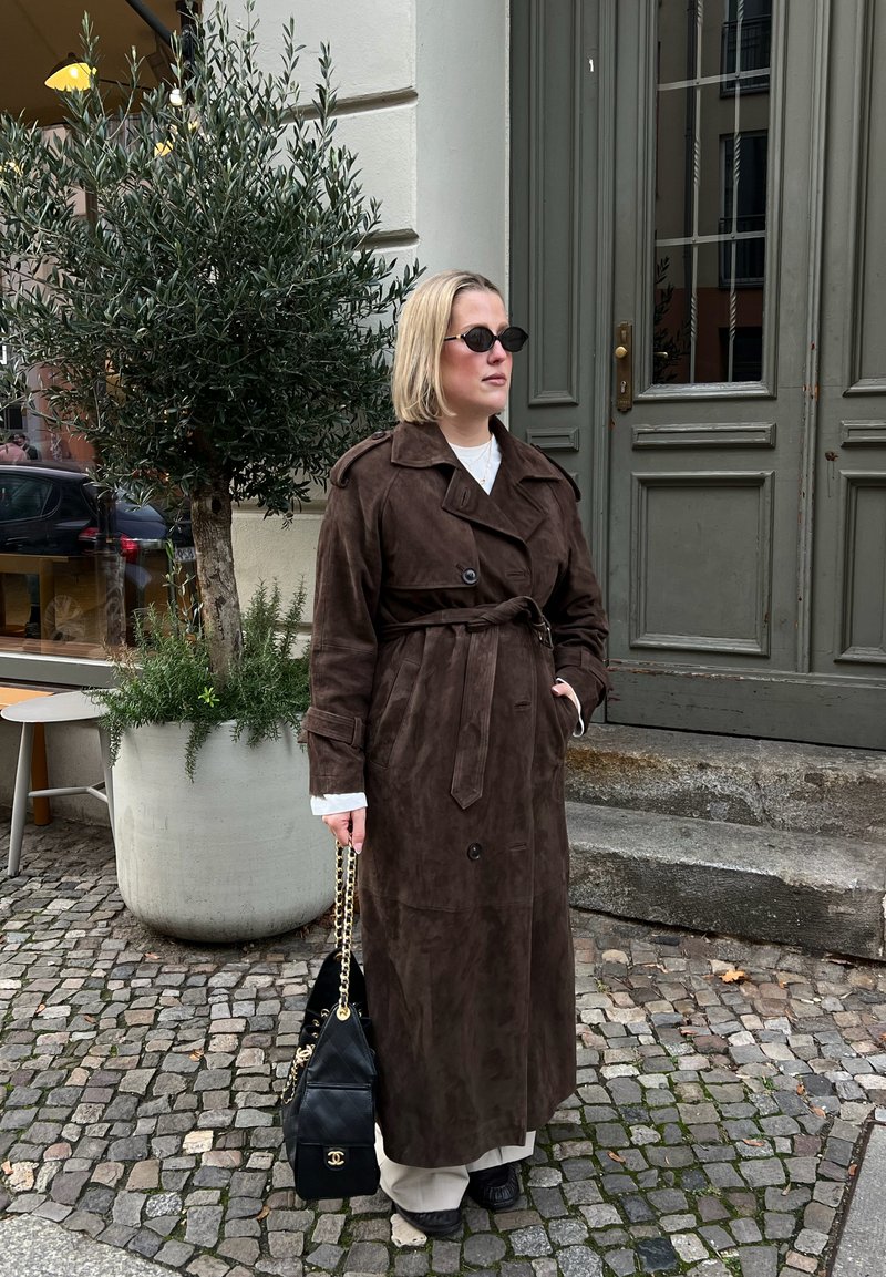Femme portant un trench-coat long marron foncé, lunettes de soleil, tenant un sac à main noir, debout dans une rue pavée près d'une grande plante en pot.