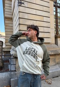 Krelles krāsas kapuce džemperis ar uzrakstu "LOCAL HEROES" tumši zaļā krāsā. Mīksts audums, priekšējā kabata un brīvs piegriezums.