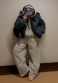 Jachetă căptușită albastru închis peste un hanorac gri; pantaloni largi de trening gri; șapcă bleumarin; căști; încălțăminte bej; chei pe un breloc.