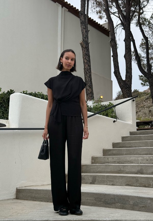 Frau in schwarzem Outfit, die auf einer Außentreppe steht und eine kleine schwarze Handtasche hält, mit weißen Wänden und Bäumen im Hintergrund.