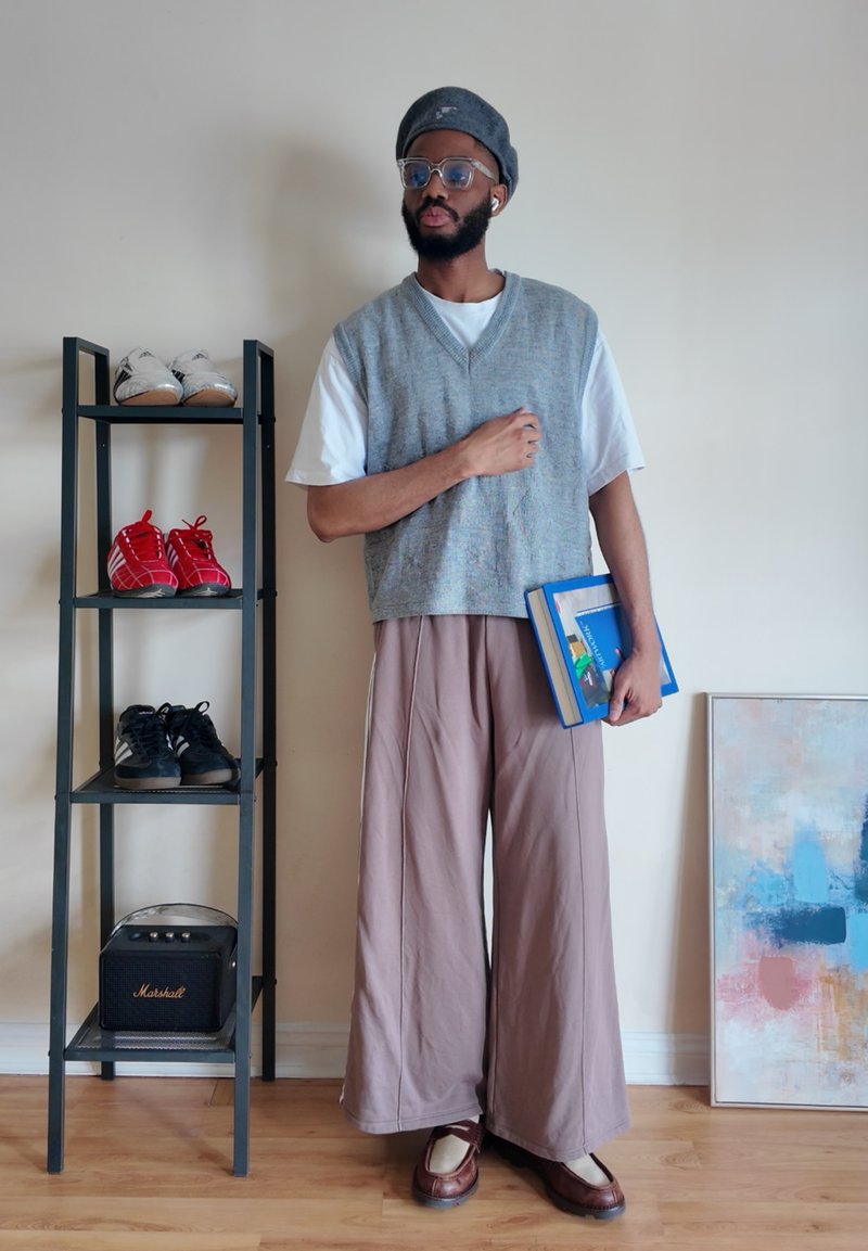 Homme portant un gilet gris et un pantalon large tenant des livres, debout à côté d'une étagère à chaussures noire et d'une peinture abstraite contre un mur beige.