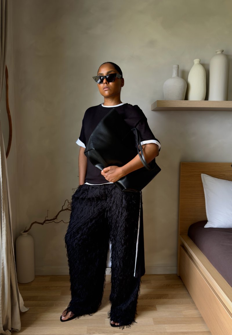 Femme portant des lunettes de soleil noires et un pantalon noir texturé, debout à l'intérieur, tenant un grand sac noir à côté du lit et des vases décoratifs.