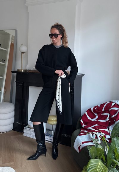 Mujer sonriendo con conjunto negro, botas hasta la rodilla y bufanda de lunares, de pie en la sala de estar cerca de la chimenea y una manta roja con patrón.