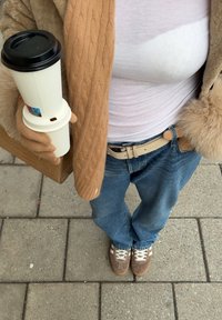 Manteau beige en tricot torsadé, haut blanc, jean bleu clair, ceinture beige, tenant une tasse de café blanche, et portant des baskets marron avec des accents blancs.