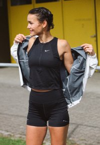 Črn športni crop top z zadrgo, parjen z visokim črnim šortsom. Zunanji sloj je siva in bela vetrovka, ki je razdrta.