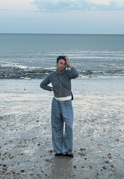 Persona con el pelo trenzado, que lleva un suéter gris y pantalones holgados, de pie en una playa rocosa y mojada con mar tranquilo y cielo nublado al fondo.