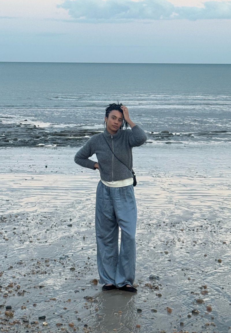 Personne aux cheveux tressés portant un pull gris et un pantalon ample se tient sur une plage rocheuse humide avec une mer calme et un ciel nuageux en arrière-plan.