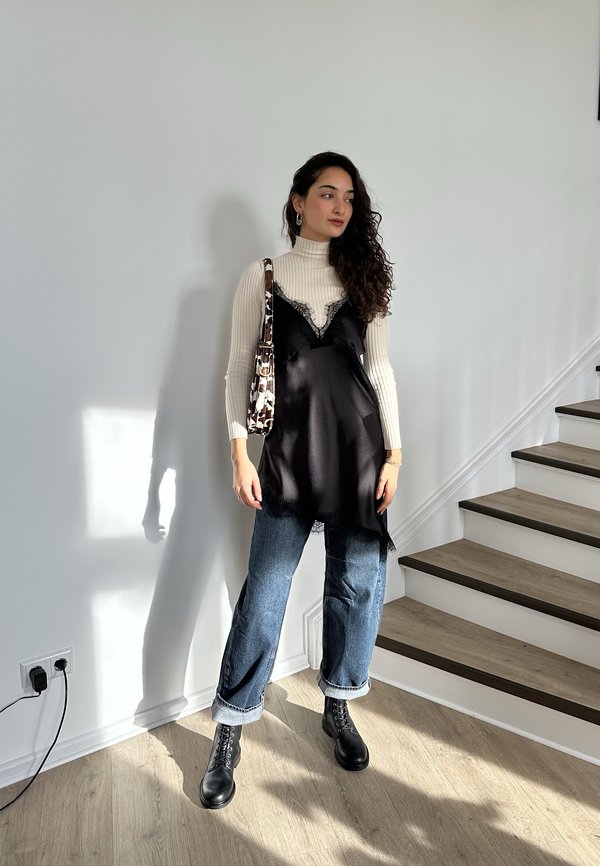 Femme aux cheveux bouclés portant un col roulé blanc, une robe en satin noir par-dessus un jean, des bottes noires, et un sac à bandoulière à motifs, à l'intérieur près des escaliers.