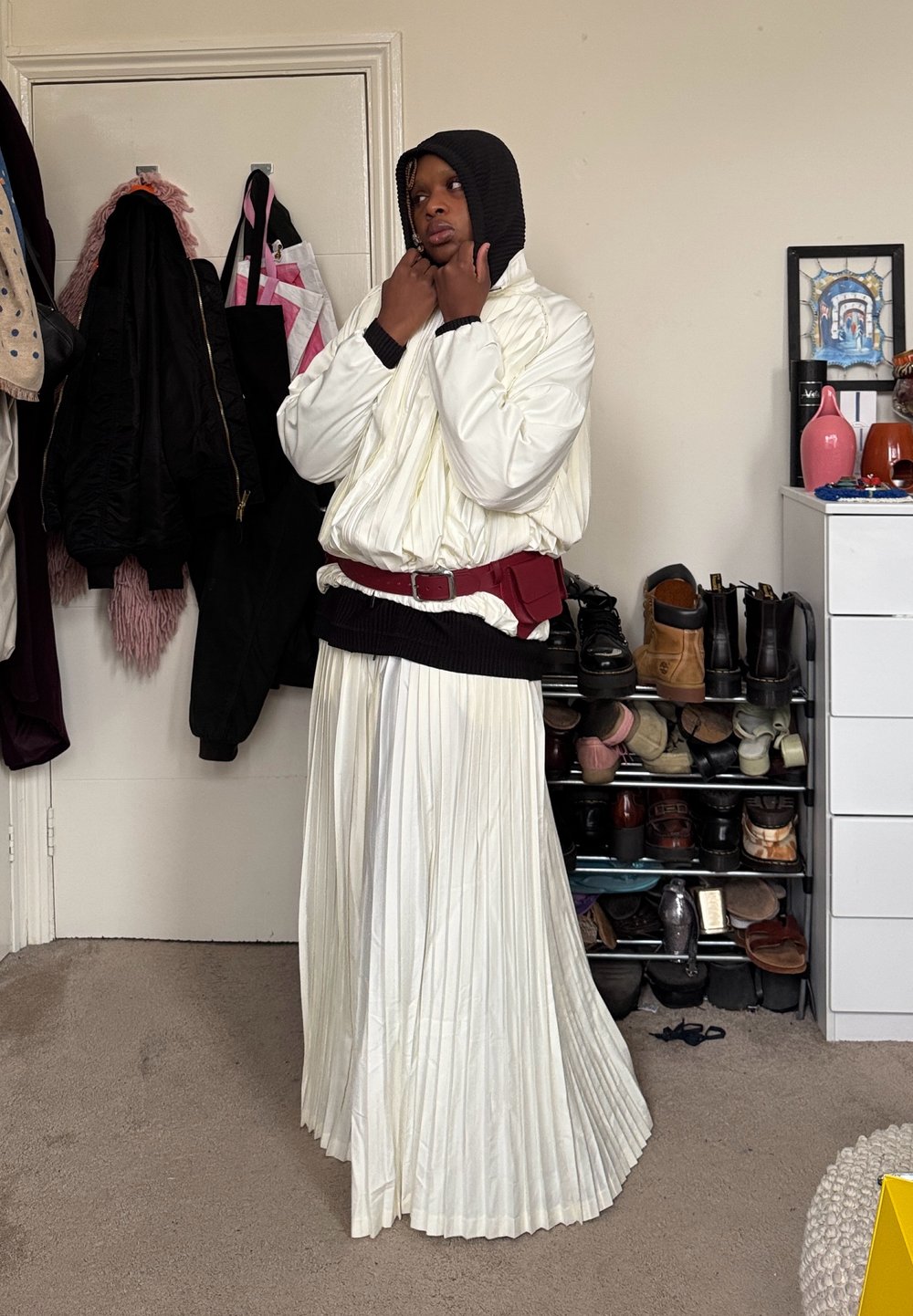 Personne portant une jupe plissée blanche, une veste bouffante blanche, une capuche noire et une ceinture-sac rouge, se tenant dans une pièce avec des manteaux et des chaussures sur des étagères.