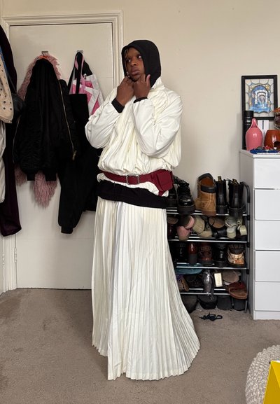 Persona con una falda blanca plisada, chaqueta blanca acolchada, capucha negra y riñonera roja, de pie en una habitación con abrigos y zapatos en estantes.