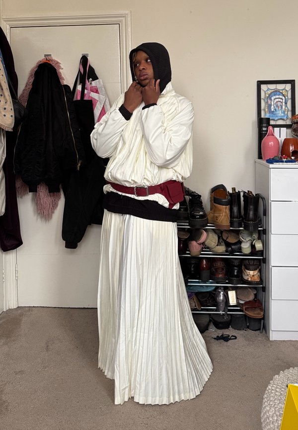 Personne portant une jupe plissée blanche, une veste bouffante blanche, une capuche noire et une ceinture-sac rouge, se tenant dans une pièce avec des manteaux et des chaussures sur des étagères.