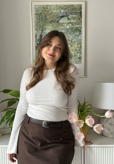 Top blanco de manga larga con ribetes, falda marrón a rayas finas con cinturón, tulipanes rosas en un jarrón, obra de arte decorativa en el fondo.