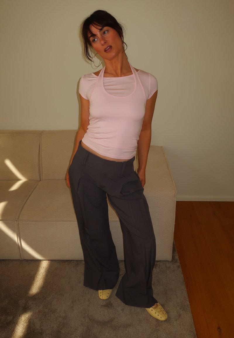Femme en top moulant rose, pantalon ample foncé et chaussures jaunes, se tient en appui vers l'arrière dans un salon beige avec des motifs de lumière du soleil sur le sol.