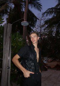 Bolsa de couro preta com várias alças, usada sobre uma camisa folgada com botões. O fundo apresenta palmeiras e uma luz suave ao entardecer.