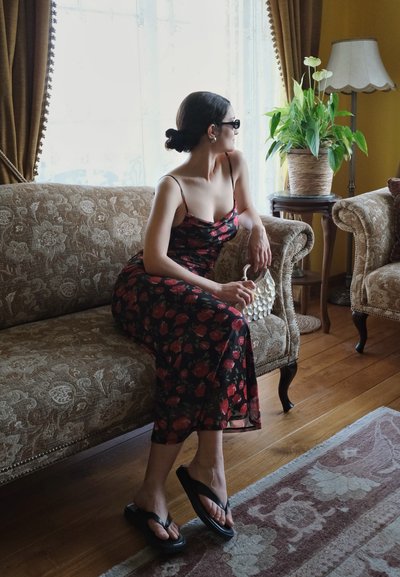 Mujer con vestido de flores y gafas de sol sentada en un sofá con motivos, sosteniendo un bolso pequeño, mirando por una ventana junto a una planta en maceta y una lámpara.