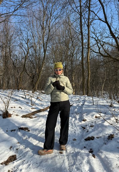 Mujer con chaqueta verde claro, pantalones negros, gorro verde y gafas de sol, de pie sobre suelo nevado en un bosque sin hojas bajo un cielo azul despejado.