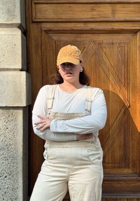 Casquette en velours côtelé beige, haut à manches longues blanc, salopette beige avec des boutons en métal, et fond en bois texturé. Position avec les bras croisés.