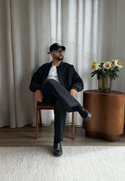 Hombre con chaqueta negra, gorra y gafas de sol sentado con las piernas cruzadas en una silla de madera junto a una mesa redonda de madera con flores.