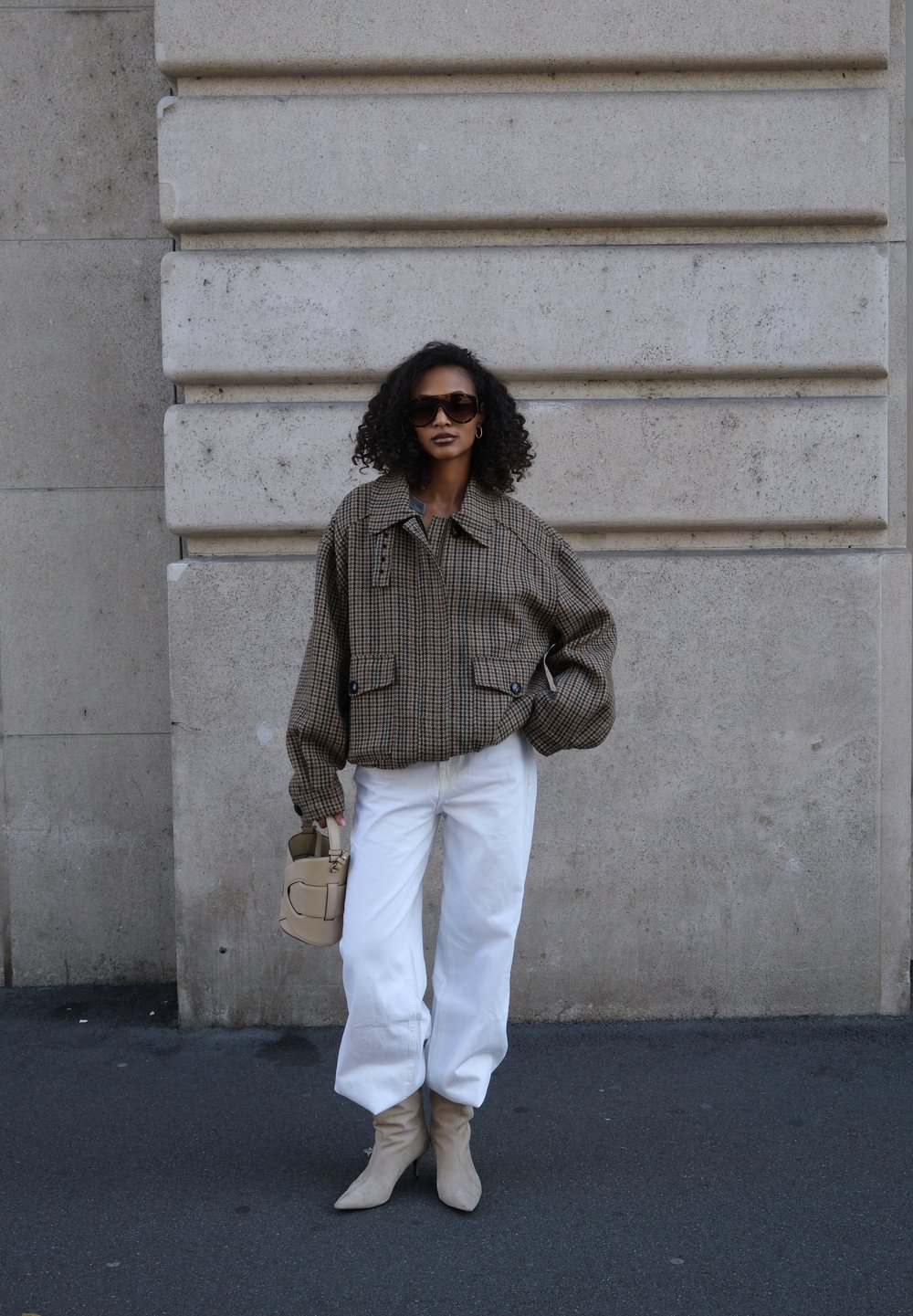 Giacca oversize marrone a quadri con due tasche anteriori, pantaloni bianchi a gamba larga, stivaletti beige con tacco e una piccola borsa beige.