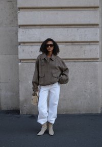 Giacca oversize marrone a quadri con due tasche anteriori, pantaloni bianchi a gamba larga, stivaletti beige con tacco e una piccola borsa beige.