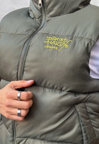 Gilet puffo verde con chiusura a zip, con ricamo frontale giallo. La texture è liscia con un motivo trapuntato. Mano che indossa anelli d'argento.