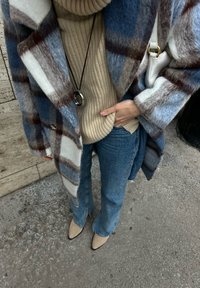 Wool karirani kaput u plavoj, bijeloj i smeđoj boji preko bež rebraste turtleneck majice; uparen s modelom traperica koje se šire prema dolje u plavoj boji i bež štiklama sa špicem.