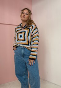 Suéter colorido de malha com um padrão geométrico de quadrados de avó nas cores rosa, azul e preto, combinado com jeans de cintura alta em azul claro.