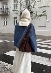 Cappello color crema in misto lana, giacca in pile blu, gonna lunga bianca e borsa in suede marrone, in piedi su una striscia pedonale sotto una leggera nevicata.