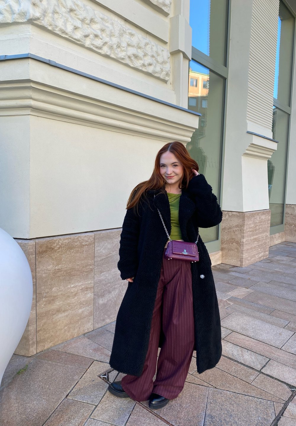 Μαύρο, αφράτο παλτό πάνω από πράσινη μπλούζα, συνδυασμένο με χαλαρούς μπορντό ριγέ παντελόνες. Μικρή μωβ τσάντα με αλυσίδα. Μαύρα επίπεδα παπούτσια.