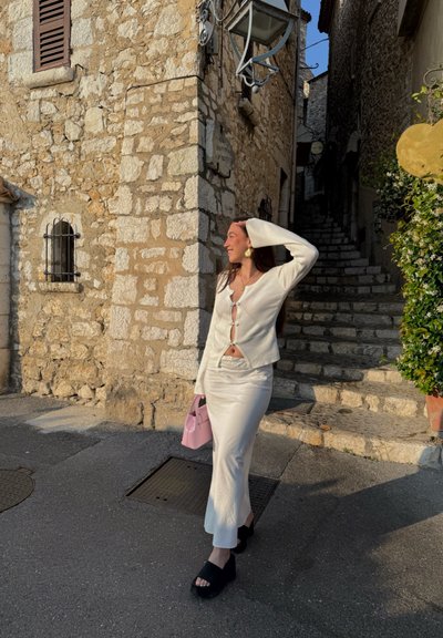 Cárdigan blanco de punto con detalles de botones, combinado con una falda blanca de satén. Lleva un pequeño bolso rosa y sandalias de cuña negras. Fondo de pared de piedra.