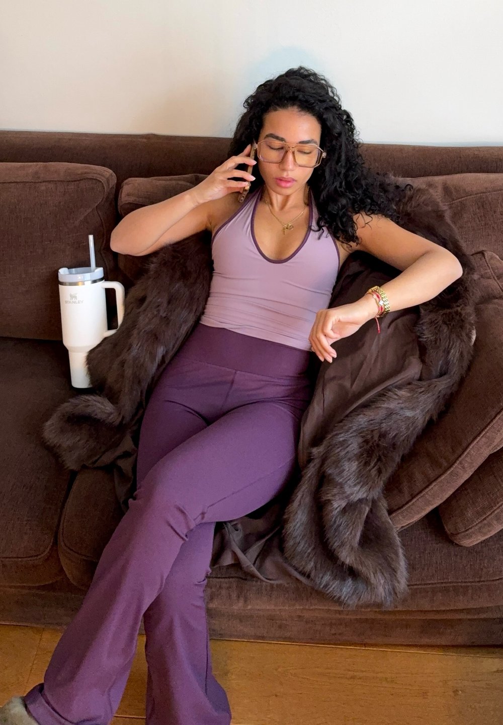 Femme en tenue de sport violette et lunettes assise sur un canapé marron, parlant au téléphone, avec un manteau en fourrure drapé et un gobelet blanc à côté d'elle.