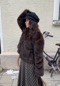 Braune Kunstpelzjacke mit langen Ärmeln, kombiniert mit einer schwarzen Mütze. Der karierte Rock sorgt für einen Musterkontrast. Im Hintergrund ist ein Fahrrad zu sehen.
