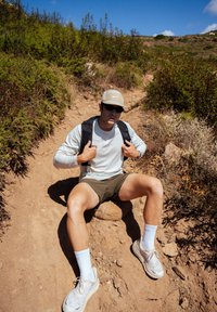 Uma pessoa sentada, vestindo uma camisa de manga longa clara e calções escuros, com uma mochila preta, óculos de sol e um boné cinza, num caminho de terra.