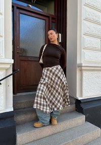 Egy illeszkedő, barna, hosszú ujjú top, amelyet egy kockás maxi szoknyával párosítanak. A farmernadrág és a barna csizmák kiegészítik az öltözetet. Háttérben egy fából készült ajtó.