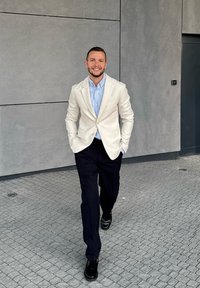 Egy világos bézs zakót, kék inget és fekete nadrágot viselő férfi sétál egy textúrált szürke burkolaton. Fekete cipőt visel és mosolyog.
