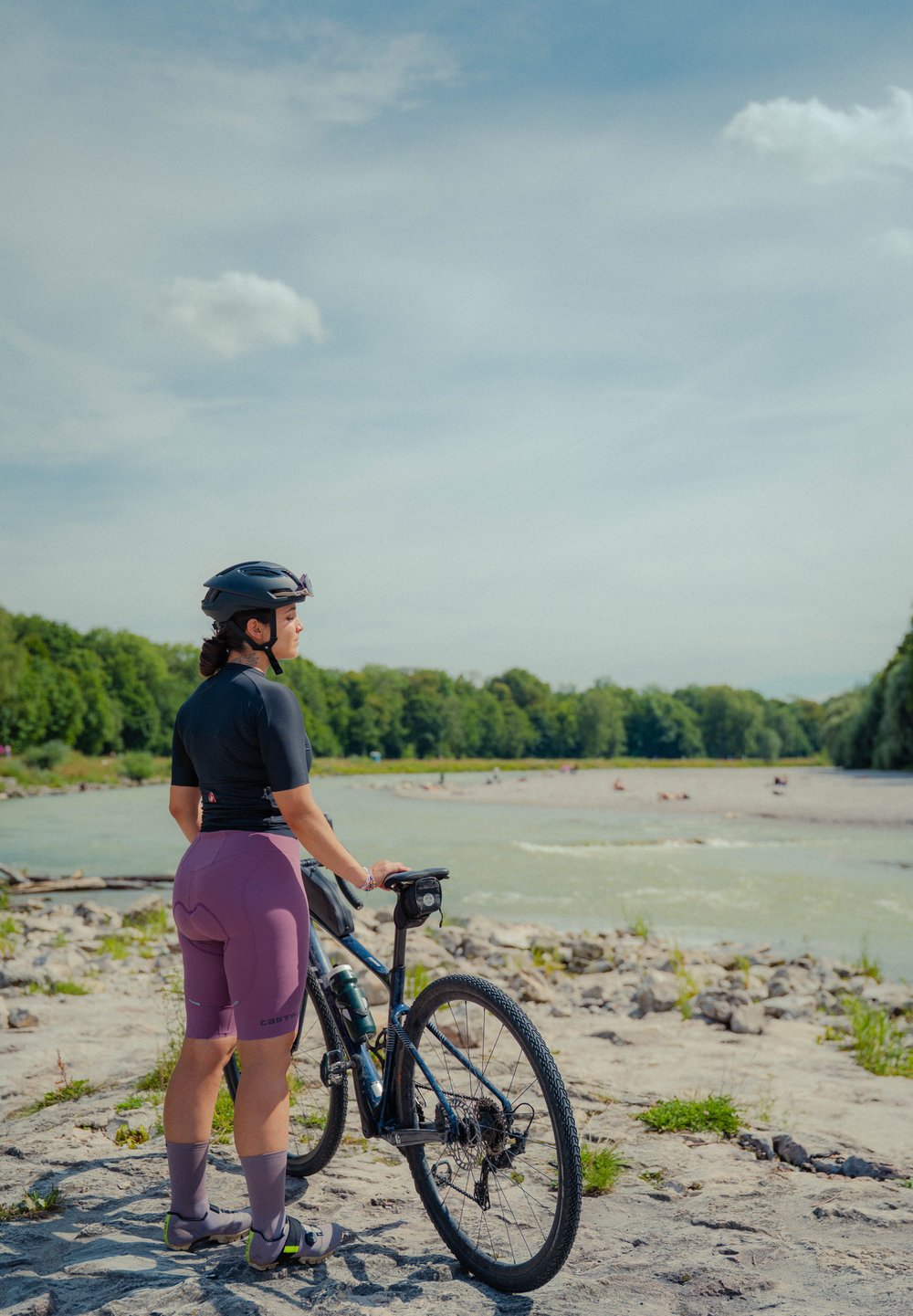 Um ciclista com uma camisola preta e calções roxos está ao lado de uma bicicleta de montanha azul em terreno rochoso, perto de um rio, com árvores ao fundo.