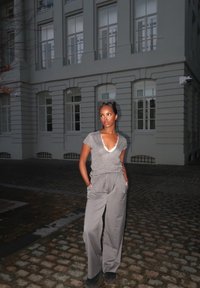 Femme debout dans une rue pavée la nuit, portant un haut gris à manches courtes et un pantalon gris ample, les mains dans les poches, regardant sur le côté.