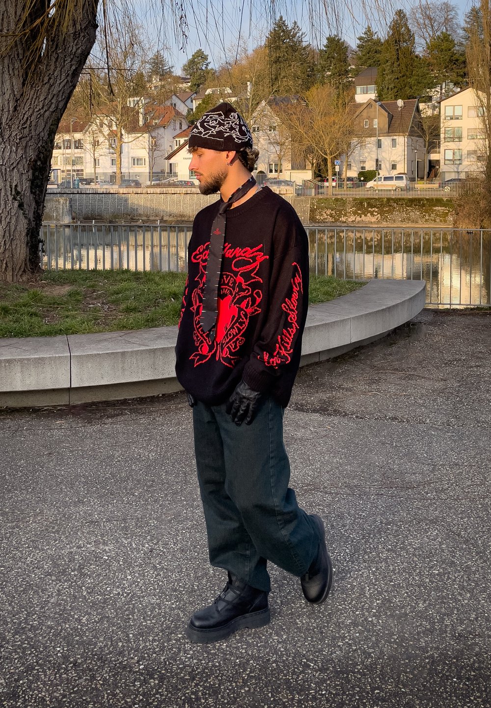 Jeune homme portant un pull noir et rouge, des gants noirs, un pantalon, des bottes et un bonnet à motif, debout près d'une rivière avec des bâtiments résidentiels en arrière-plan.