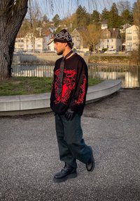 Jeune homme portant un pull noir et rouge, des gants noirs, un pantalon, des bottes et un bonnet à motif, debout près d'une rivière avec des bâtiments résidentiels en arrière-plan.