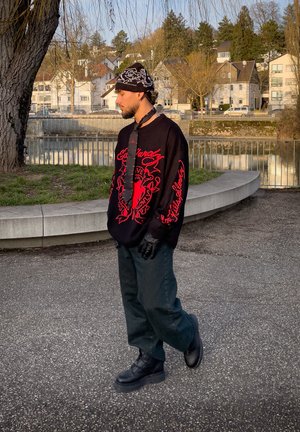 Joven con suéter negro y rojo, guantes negros, pantalones, botas y gorro estampado, de pie junto al río con edificios residenciales al fondo.