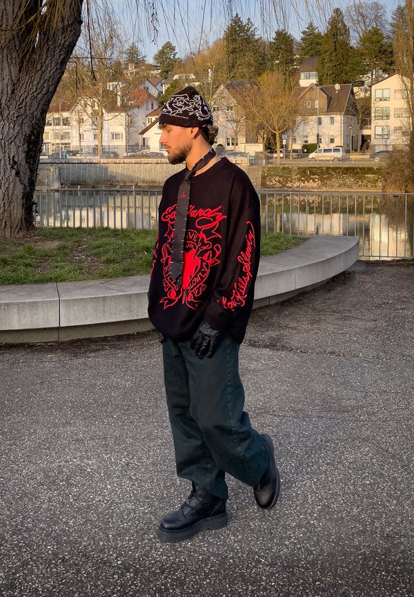 Jonge man die een zwart-rode trui, zwarte handschoenen, een broek, laarzen en een gebreide muts met patroon draagt, staand bij een rivier met woongebouwen op de achtergrond.