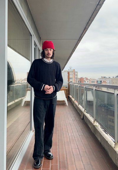 Suéter negro de punto trenzado con detalles blancos, pantalones anchos negros y zapatos negros. Gorra roja, de pie en un balcón con vista a la ciudad.