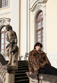 Jeune homme en manteau de fourrure à imprimé léopard assis sur un rebord en pierre à côté d'une statue classique près des marches d'un grand bâtiment aux fenêtres arquées.