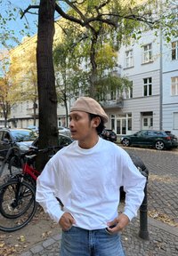 Weißes Langarmhemd, blaue Jeans und beigefarbene Baskenmütze. Draußen mit Bäumen und geparkten Autos im Hintergrund. Lässige und entspannte Pose.